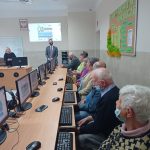 Zdjęcie przedstawia osoby w wieku od 60 do 80 lat przed komputerami. W oddali stoi Wójt Gminy Wietrzychowice oraz Pani prowadząca zajęcia komputerowe dla seniorów.