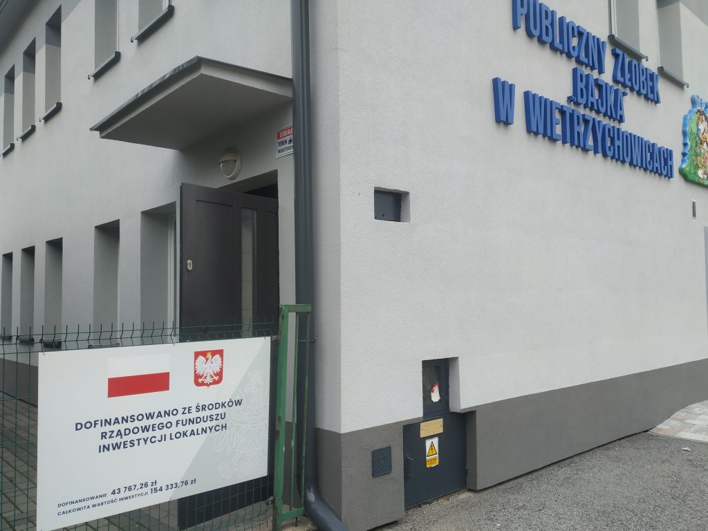 Zdjęcie przedstawia fragment bdynku od strony wejścia do Publicznego Żłobka "Bajka" w Wietrzychowicach oraz tablicę informacyjną o dofinnasowaniu zadania.