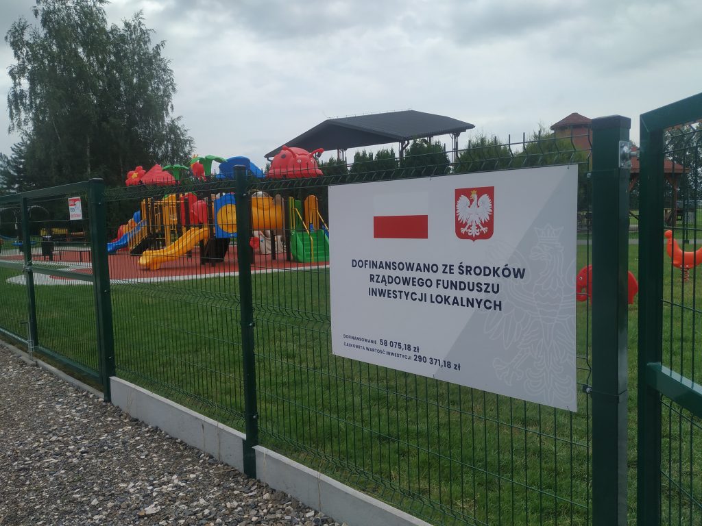 Zdjęcie przedstawia tablicę informacyjną zawieszoną na ogrodzeniu z informacją o dofinansowaniu zadania, w tle plac zabaw w Wietrzychowicach obok przedskzola.