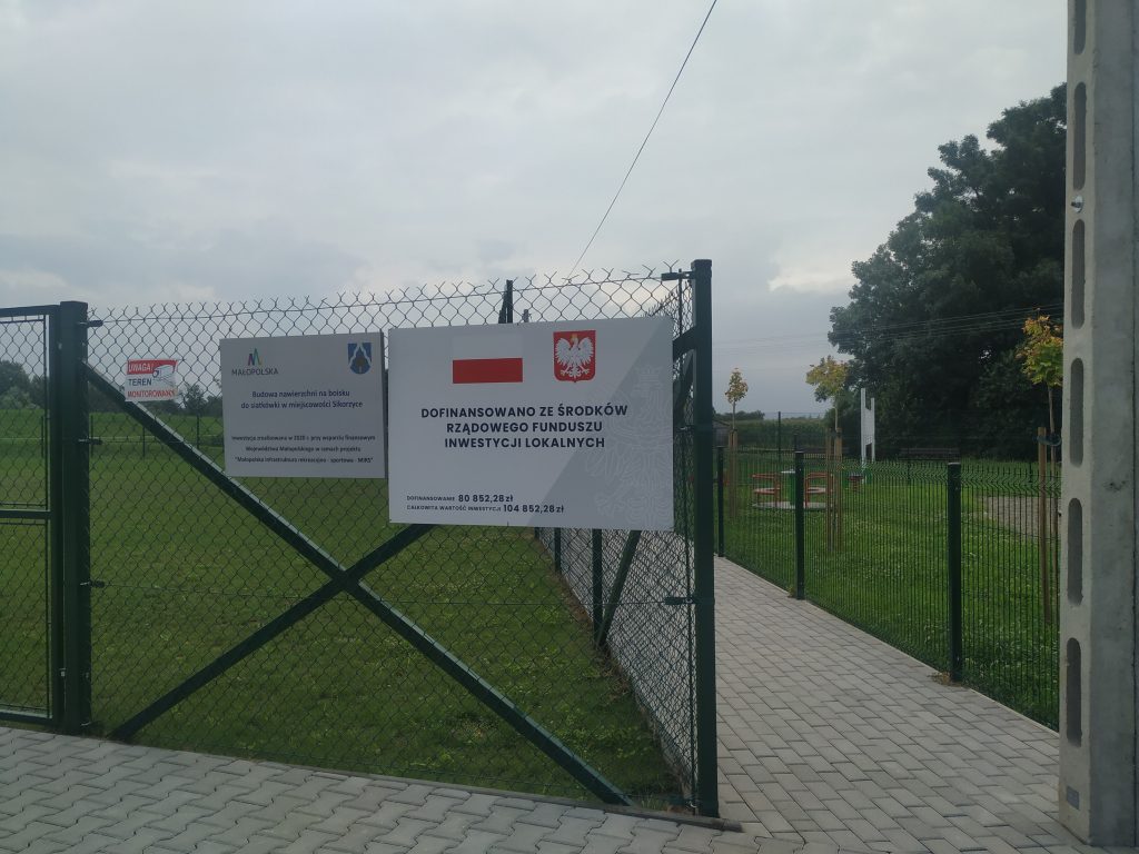 Zdjęcie przedstawia tablice informacyjną o dofinansowaniu zadania budowy nawierzchni na boisku do siatkówki w miejscowosci Sikorzyce.