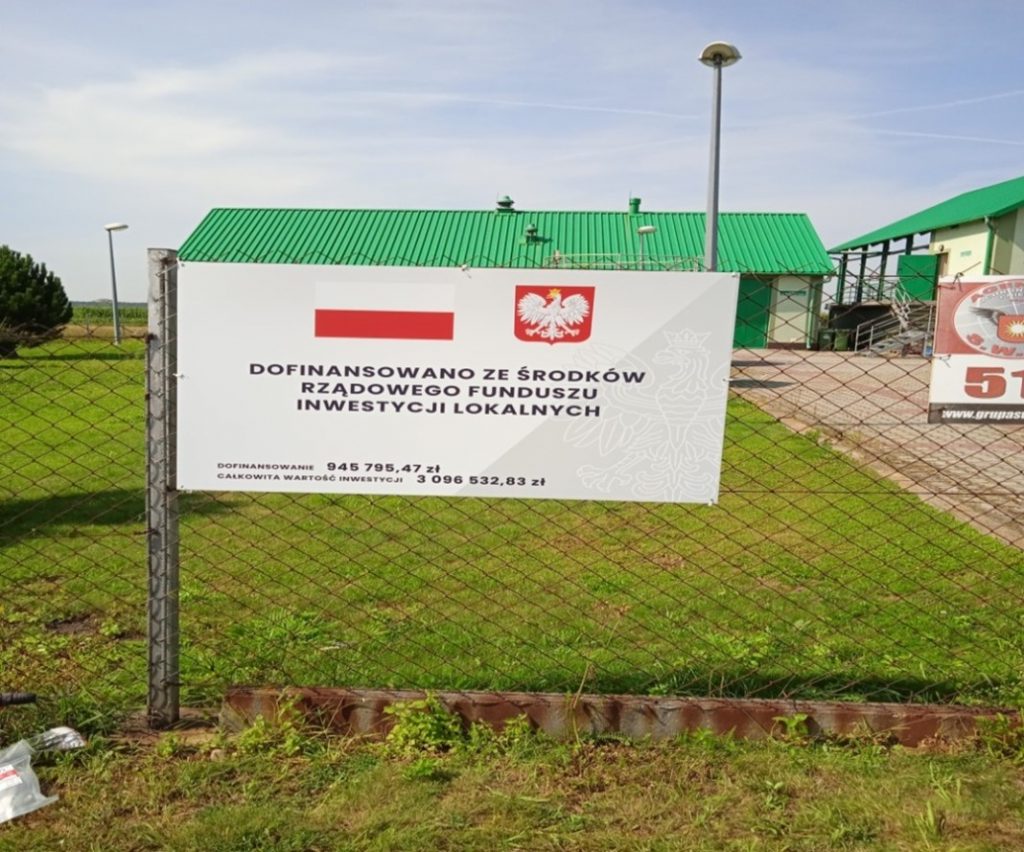 Zdjęcie przedstawia tablice informacyjną o dofinansowaniu zadania budowy sieci kanalizacji sanitarnej w miejscowosci Jadowniki Mokre, w tle budynki Zakładu oczyszczalni ścieków w Miechowicach Wielkich.